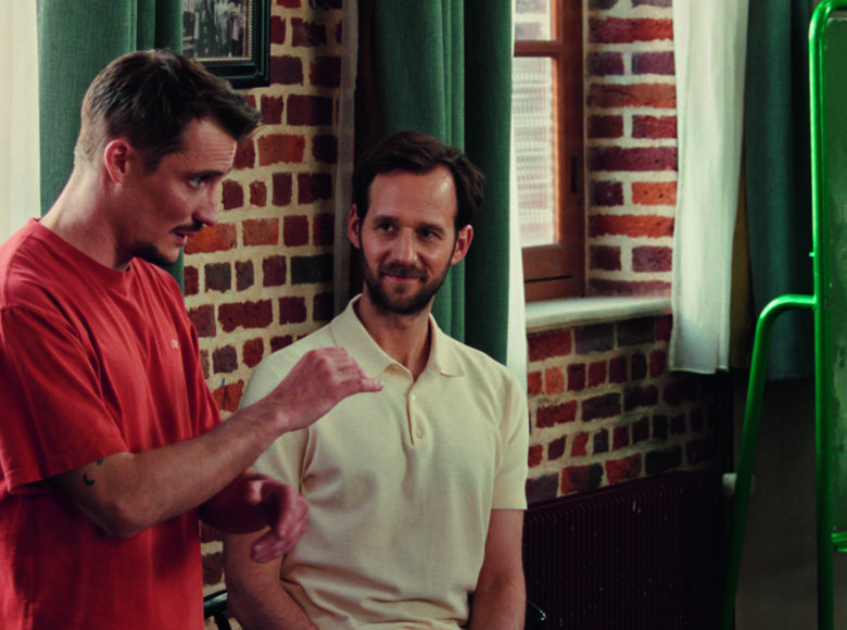 Kadr z filmu. W pomieszczeniu pod ścianą z oknami stoją dwaj mężczyźni, rozmawiając ze sobą. Obok nich stoi tablica szkolna, na której narysowane są pięciolinie i inne oznaczenia muzyczne.