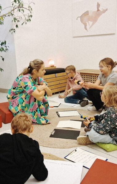 W pomieszczeniu kilkoro dzieci z opiekunką, siedzące w kręgu na podłodze na poduszkach. Przed dziećmi leżą kartki i kredki. Niektóre z dzieci trzymają w rękach lalki.