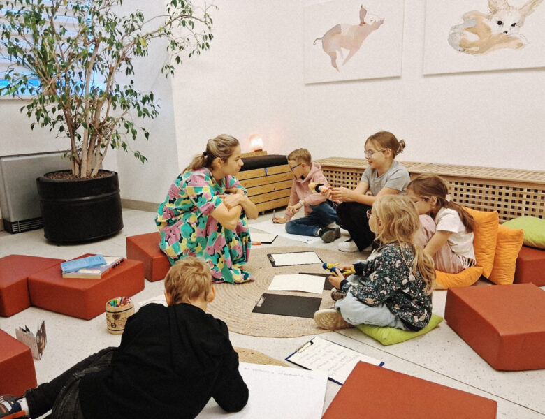 W pomieszczeniu kilkoro dzieci z opiekunką, siedzące w kręgu na podłodze na poduszkach. Przed dziećmi leżą kartki i kredki. Niektóre z dzieci trzymają w rękach lalki.