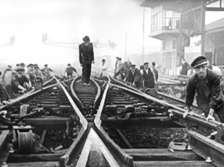 Kadr z filmu czarno biały. Czasy przedwojenne. Duża grupa mężczyzn układających tory kolejowe.