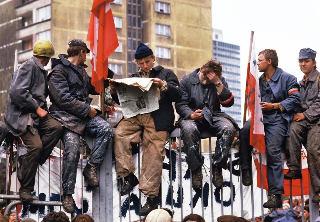 Na kolorowej fotografii dokumentującej strajk w Stoczni Gdańskiej, widzimy grupę 6 siedzących na płocie Stoczni robotników, ubranych w zniszczone stroje robocze. Dwóch ma na prawej ręce biało-czerwoną opaskę, jeden czyta rozłożoną gazetę, inny trzyma biało-czerwoną flagę. Na drugim planie widać zawieszony na płocie Stoczni napis na białym płótnie „STRAJK OKUPACYJNY”