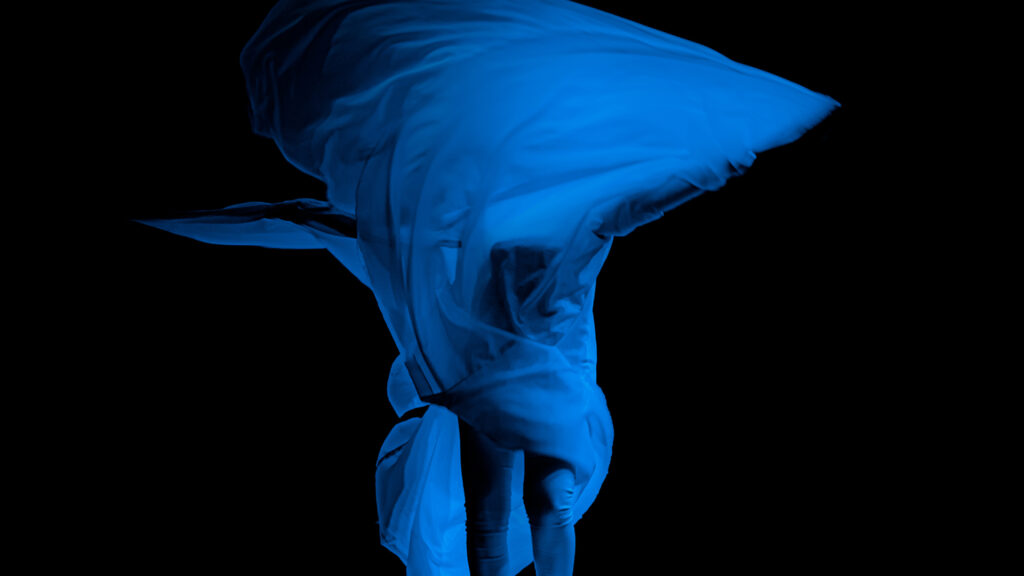 Na kolorowej fotografii, na czarnym tle, widzimy uchwyconą w ruchu kręcącą się w tańcu?postać, owiniętą w białą tkaninę, która jest podświetlona niebieskim światłem.