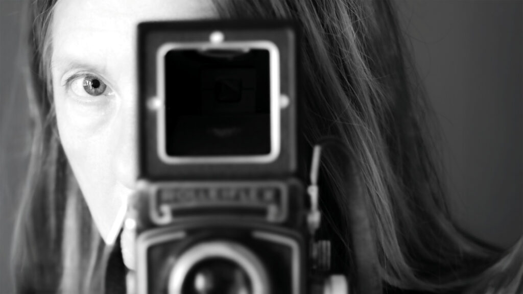 Fotografia czarno biała. Duże zbliżenie na twarz kobiety patrzącej w obiektyw starego, analogowego aparatu fotograficznego. Aparat ustawiony jest na wprost i zasłania większość twarzy kobiety tak, że widzimy tylko jedno jej oko oraz włosy.