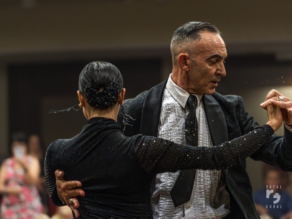 Na kolorowej fotografii, widzimy parę tańczącą tango, w planie od pasa w górę. Ubrana na czarno kobieta jest odwrócona do nas tyłem, ma czarne, spięte czarną kokardą włosy. Mężczyzna jest widoczny z przodu. To brunet z klasyczną, krótką fryzurą, jest ubrany w ciemną marynarkę, jasną koszulę i ciemny krawat. Prowadząc tancerkę ma rozłożone ramiona w pozycji klasycznej ramy. Ich dłonie są splecione. Jergo twarz jest zwrócona profilem w kierunku tańca.