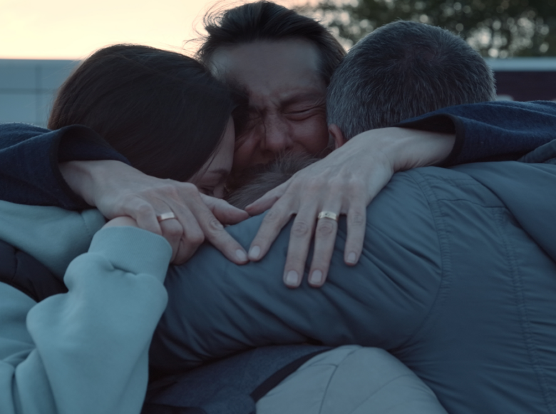 Kadr z filmu. Zbliżenie na trzy obejmujące się osoby. W tle zabudowania i drzewa.