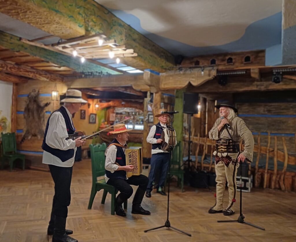 Wnętrze karczmy wiejskiej. Na środku czterech mężczyzn w strojach ludowych. jeden z nich śpiewa do mikrofonu, pozostali grają na akordeonie, skrzypcach i bębnie.