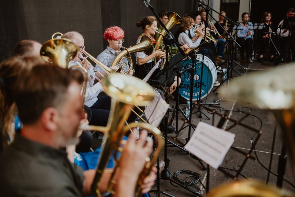 W dużym pomieszczeniu na krzesłach obok siebie w rzędzie siedzi kilkanaście osób, grających na instrumentach dętych.