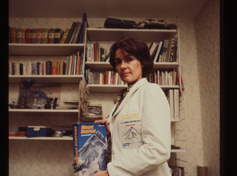 Kadr z filmu. W pokoju, na tle półek z książkami stoi kobieta ubrana w biały fartuch. W rękach trzyma książkę pod tytułem: "Mount Everest". Na fartuchu ma umieszczony napis po angielsku: "Mount Everest Expedition '78. Wanda Rutkiewicz. Poland" i logo Polskich Linii Lotniczych LOT.