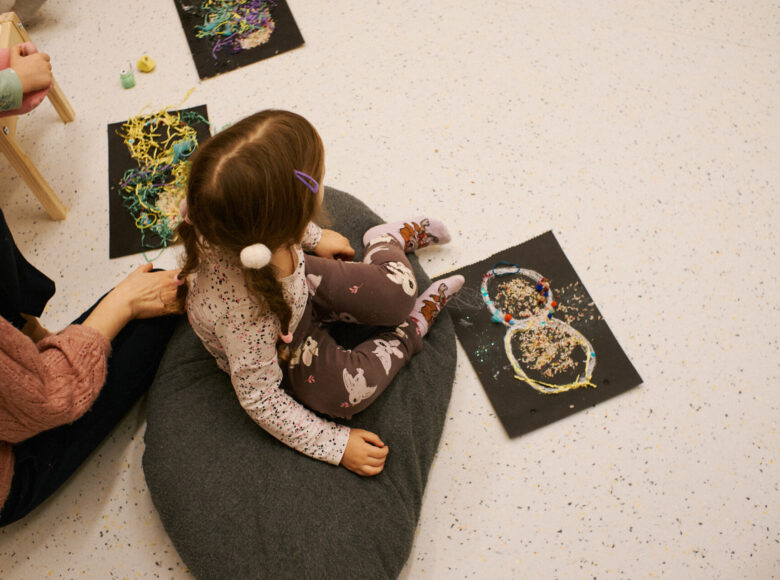 Sfotografowana od góry dziewczynka, siedząca na podłodze na dużej poduszce. Przed dziewczynką na podłodze leżą materiały do robienia ozdób.