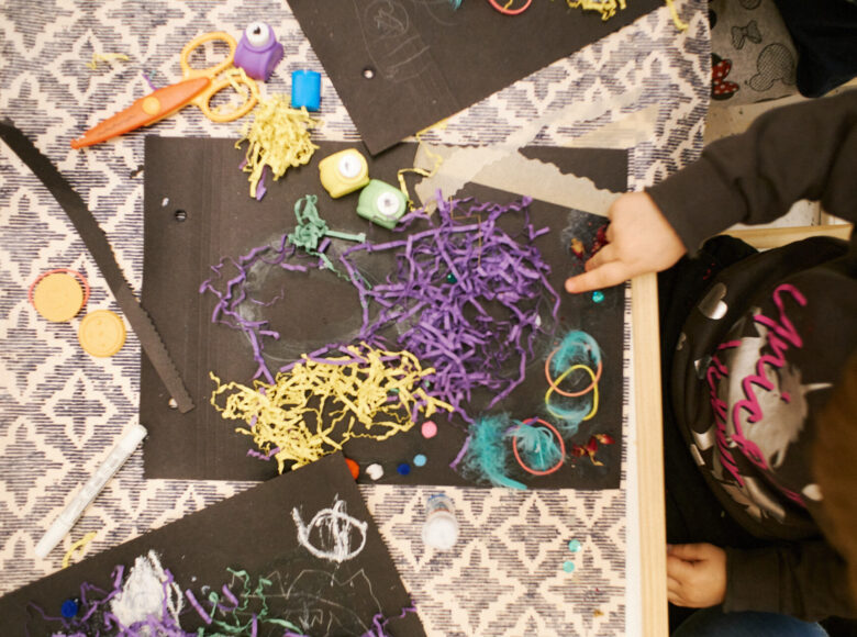 Sfotografowany od góry stół, na którym leżą materiały papiernicze do tworzenia ozdób. Po materiały sięga ręka kilkuletniego dziecka, które znajduje się poza kadrem.