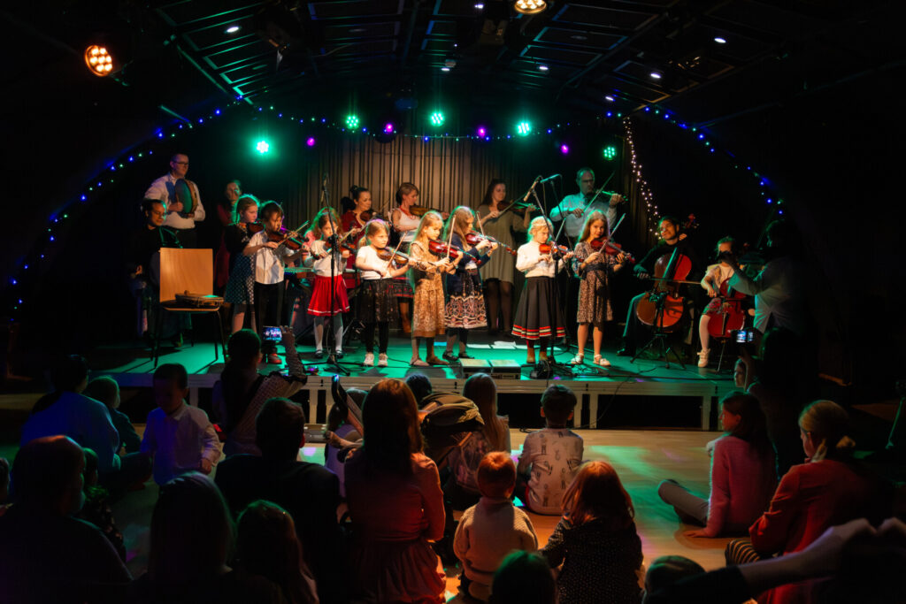 Na scenie stoi grupa złożona z dziewczynek, jednego chłopca oraz kilku dorosłych. Osoby te grają na skrzypcach, wiolonczelach i tamburynie. Przed sceną siedzą na podłodze dzieci, oglądające występ.