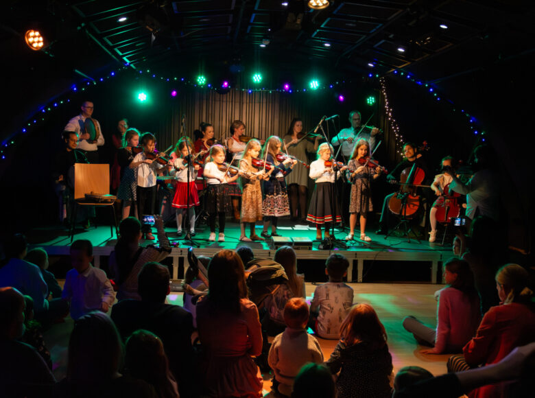 Na scenie stoi grupa złożona z dziewczynek, jednego chłopca oraz kilku dorosłych. Osoby te grają na skrzypcach, wiolonczelach i tamburynie. Przed sceną siedzą na podłodze dzieci, oglądające występ.