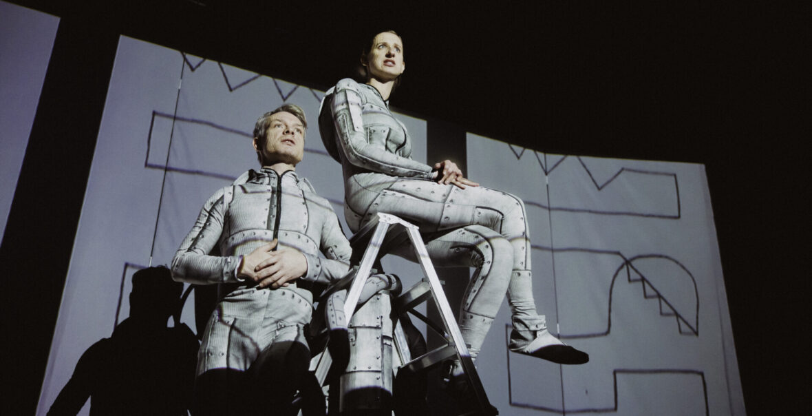 Przedstawienie teatralne. Na scenie aktorka i aktor ubrani w szare kombinezony. Aktorka niedzi na aluminiowej drabinie stojącej pośrodku sceny. Obok stoi aktor, opierając się łokciem na szczeblu drabiny. Za aktorami dekoracja złożona z wiszacych białych płacht pokrytych rysunkami przedstawiającymi kształty geometryczne. Z tyłu czarne tło.