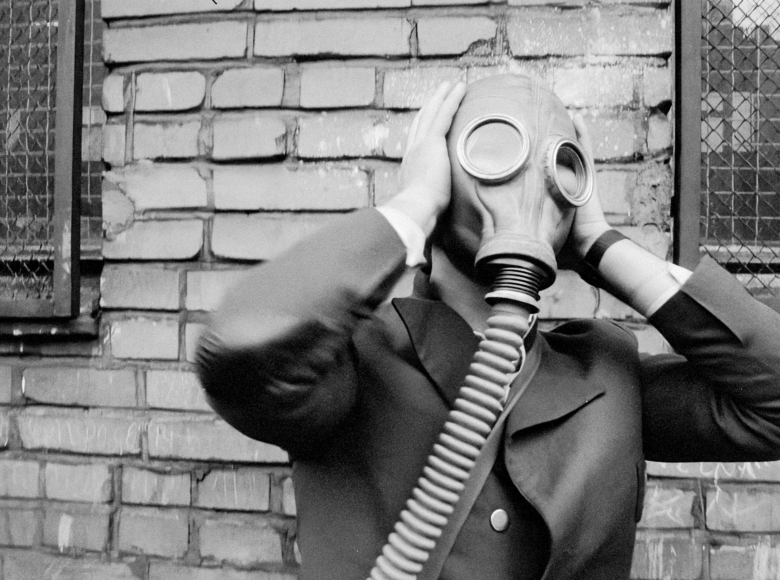 Kadr z filmu czarno biały. Na tle ceglanego budynku stoi mężczyzna w płaszczu i masce gazowej na głowie.