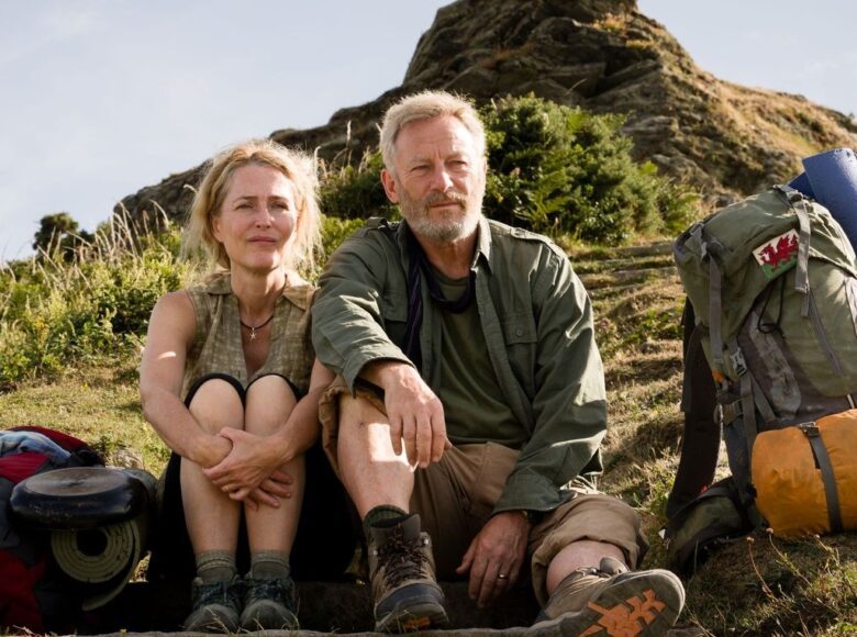 Kadr z filmu. Na trawie siedzą obok siebie kobieta i mężczyzna, oboje w średnim wieku. Obok nich leżą dwa plecaki turystyczne. W tle za nimi widzimy skałę i jasne niebo.