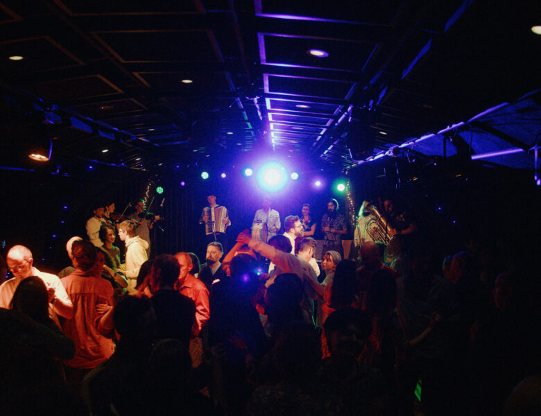W sali na pierwszym planie gęsty tłum tańczących ludzi. Na dalszym planie scena, na której grupa osób gra na instrumentach muzycznych. Sala i scena oświetlone są punktowym światłem reflektorów.