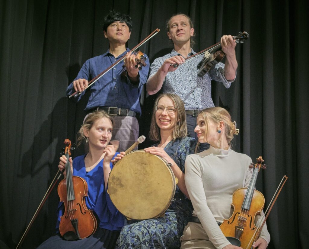 Na tle czarnej kurtyny do fotografii pozuje grupa osób - trzy kobiety i dwóch mężczyzn. jedna z kobiet trzyma w ręku bęben, pozostali trzymają skrzypce.