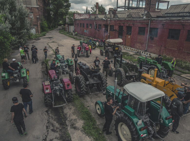 Kadr z filmu. Na placu przed przemysłowym budynkiem z cegłu stoi kilka- kilkanaście traktorów. Między traktorami chodzą ludzie.