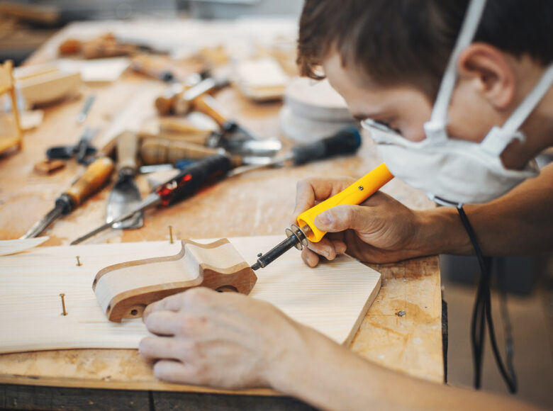 Przy stole warsztatowym siedzi mężczyzna i rzeźbi dłutkiem drewniany ornament wyglądający jak fragment mebla.
