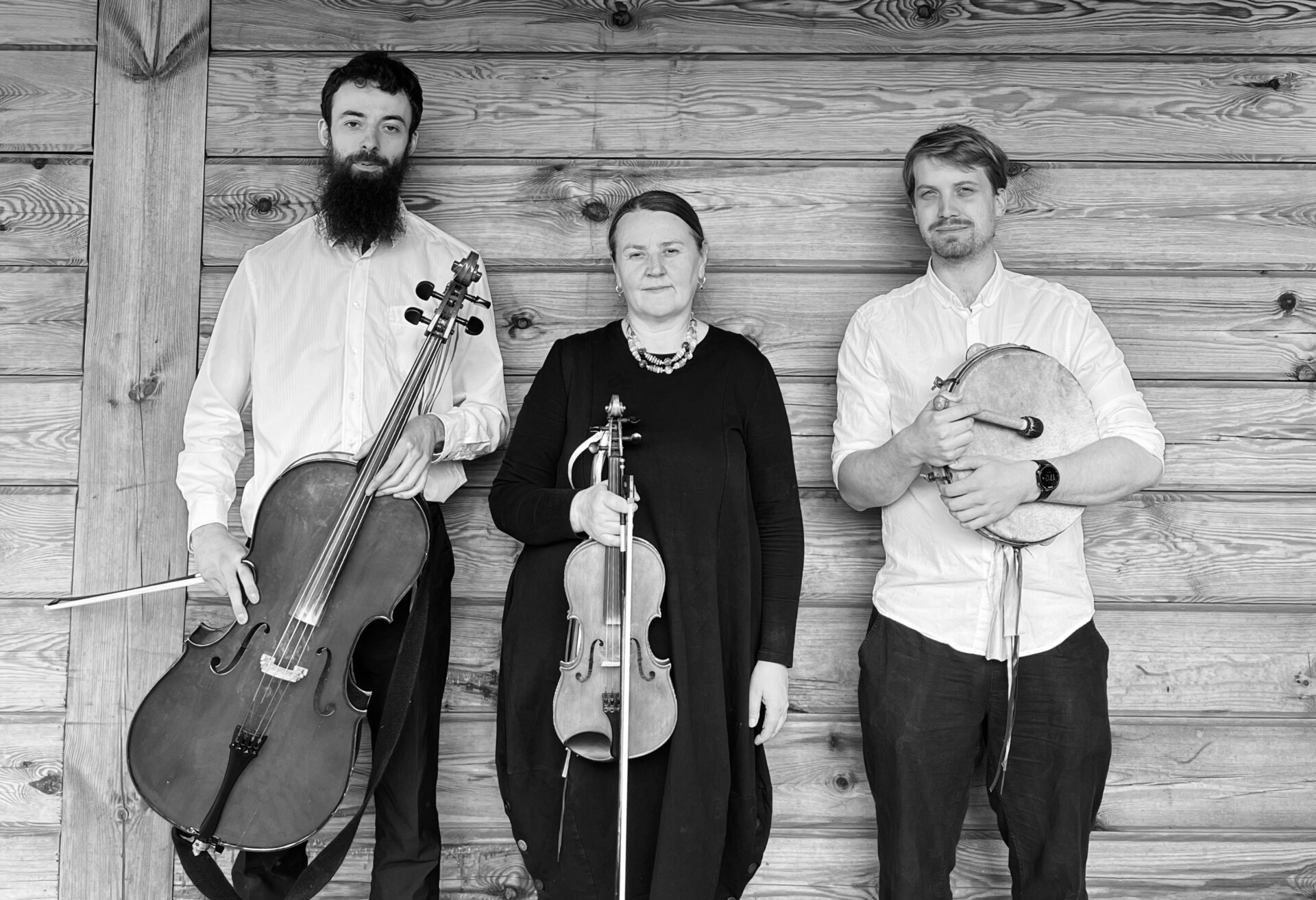 Fotografia czarno biała. Na tle drewnianej ściany wiejskiego domu stoi kobieta w średnim wieku i dwóch młodych mężczyzn. Kobieta trzyma w ręku skrzypce. Jeden z mężczyzn trzyma wiolonczelę. Drugi trzyma tamburyn.