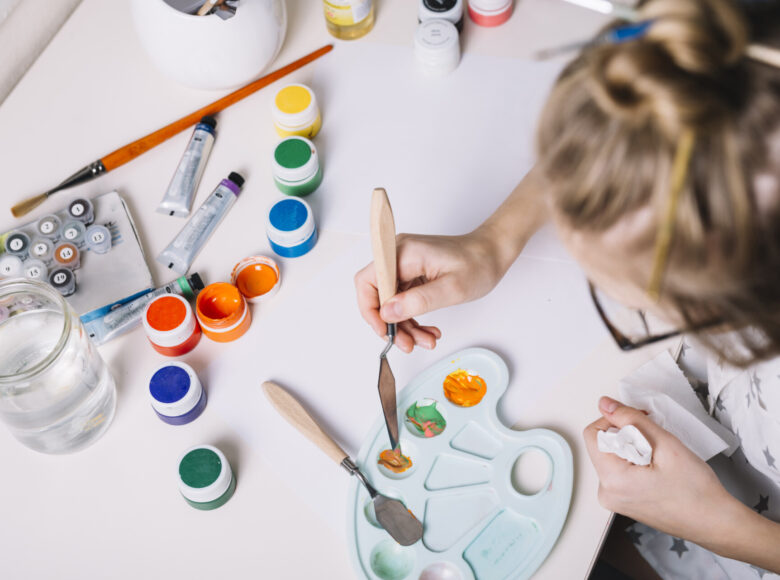 Zdjęcie zrobione od góry. Zbliżenie na głowę i ręce dziewczynki siedzącej przy stole i mieszającej szpachelką farby na palecie malarskiej.