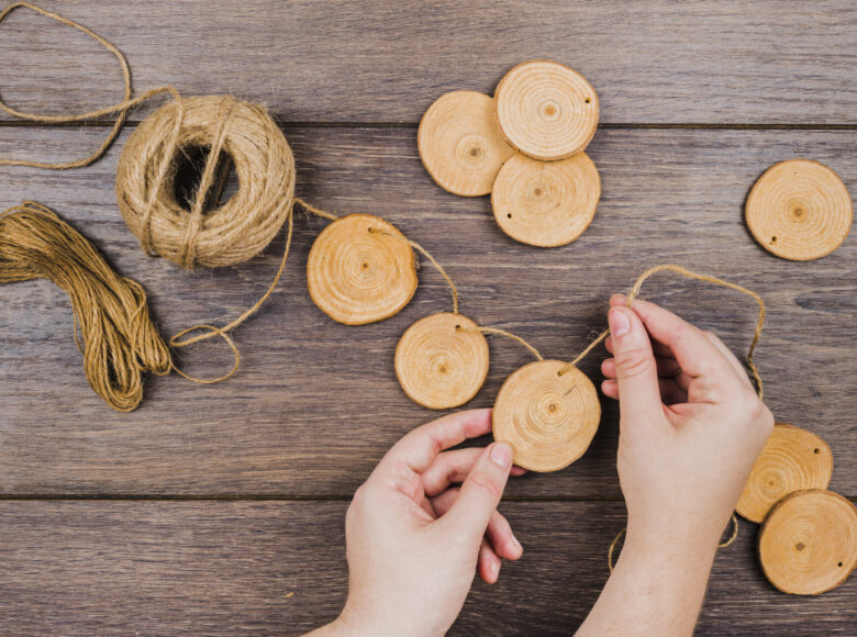 Zdjęcie zrobione od góry. Zbliżenie na dłonie młodej osoby, oparte na drewnianym blacie stołu. Dłonie trzymają lniany sznurek, na który nawleczone są okrągłe, płaskie kawałki drewna.