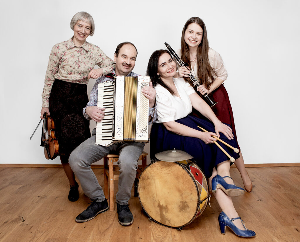 Na tle białej ściany cztery osoby - trzy kobiety i mężczyzna - pozują do fotografii. Osoby te trzymają w rękach różne instrumenty muzyczne - skrzypce, klarnet, akordeon oraz pałeczki od bębna, który stoi obok.