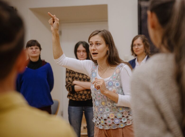 W sali stoi kobieta, która tłumaczy coś zgromadzonym wokół niej osobom.
