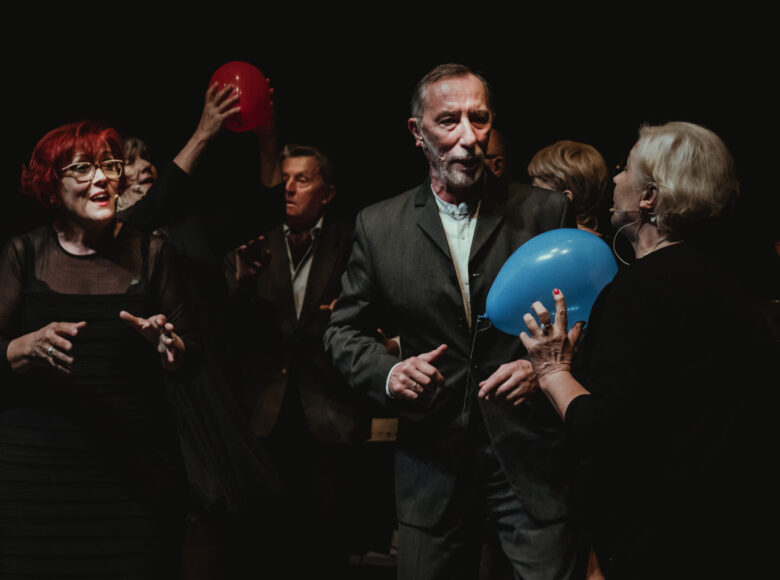 Przedstawienie kabaretu. Na scenie grupa starych osób, kobiet i mężczyzn, stojących blisko obok siebie. Dwie z kobiet trzymają w dłoniach balony i podają je do stojących obok mężczyzn.