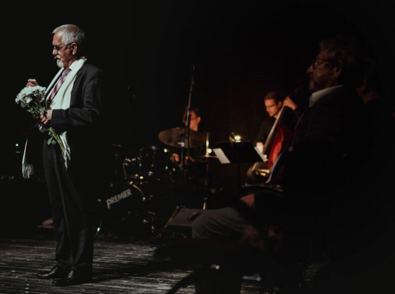Na scenie oświetlonej skąpym światłem stoi starszy mężczyzna w czarnym garniturze, trzymając w ręku bukiet białych kwiatów. Za nim siedzi grupa muzyków z instrumentami muzycznymi.