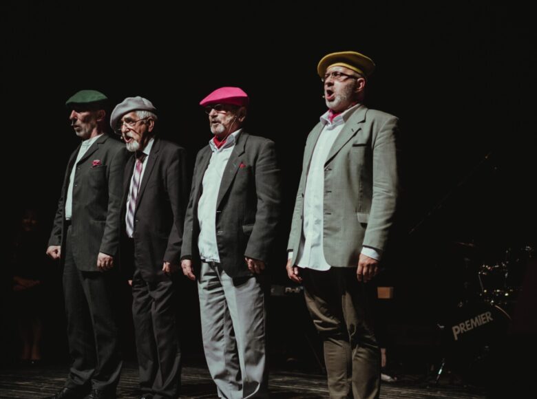 Na scenie czterej starzy mężczyźni stojący w rzędzie i śpiewający. Ubrani są w marynarki i spodnie oraz berety. Za nimi w półmroku grupa muzyków siedzących przy instrumentach.