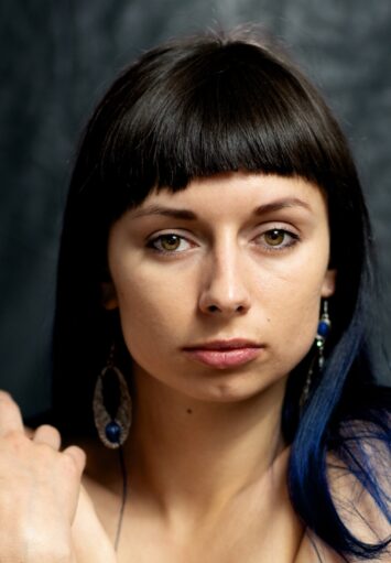Zbliżenie na twarz młodej kobiety o długich, ciemnych włosach, w delikatnym makijażu i dużych kolczykach. Kobieta patrzy prosto w obiektyw. Za kobietą niedookreślone, ciemne tło.