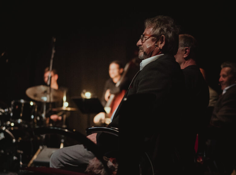 Na scenie oświetlonej skąpym światłem siedzi grupa muzyków przy instrumentach muzycznych.