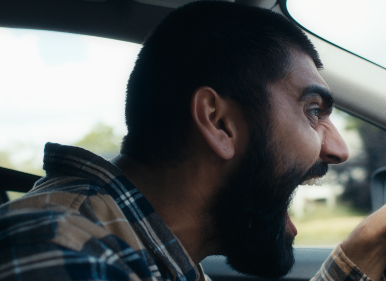 Wnętrze samochodu. Zbliżenie na twarz i ulożone na kierownicy ręce mężczyzny kierującego samochodem. Mężcxyzna nachyla się na kierownicą i patrzy przed siebie otwierając usta. Wygląda, jakby krzyczał.