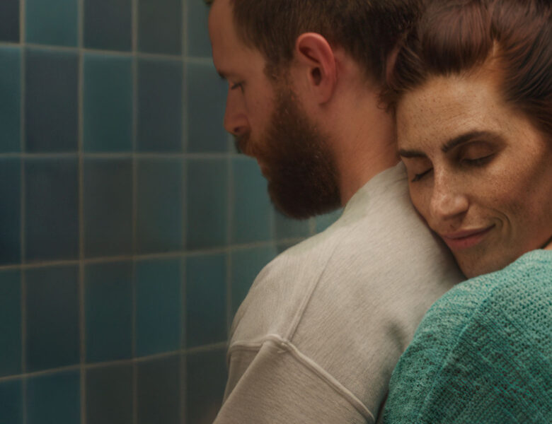 Zbliżenie na młodego mężczyzną i obejmującą go od tyłu młodą kobietę. Za nimi ściana obłożona niebieskimi kafelkami.