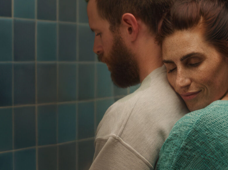 Zbliżenie na młodego mężczyzną i obejmującą go od tyłu młodą kobietę. Za nimi ściana obłożona niebieskimi kafelkami.