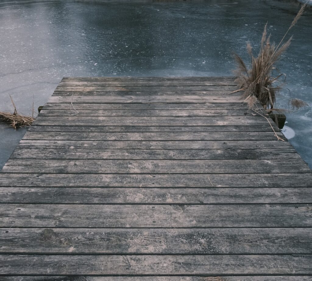 Kolorowa fotografia przedstawia koniec pomostu, zbudowanego ze starych już szarych desek, na tle zamarzniętej wody. Po bokach pomostu widać po obu stronach małe kępy wodnych traw.