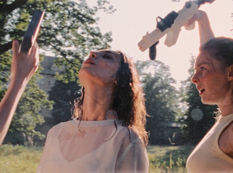 Słoneczny letni dzień, na dworze. Zbliżenie na dwie młode kobiety stojące obok siebie. Obie mają mokre włosy. Jedna z kobiet unosi do góry rękę, trzymając w niej pistolet na wodę. Obok widoczna podniesiona do góry ręka kobiety znajdującej się poza kadrem; ta ręka również trzyma pistolet na wodę. W tle trawa i zielone drzewa, a za nimi dom jednorodzinny.