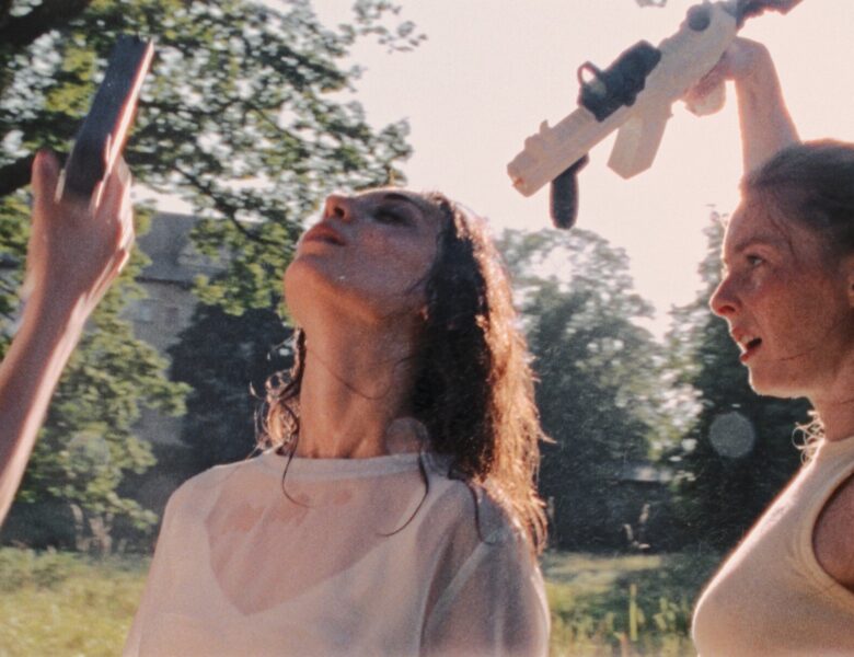 Słoneczny letni dzień, na dworze. Zbliżenie na dwie młode kobiety stojące obok siebie. Obie mają mokre włosy. Jedna z kobiet unosi do góry rękę, trzymając w niej pistolet na wodę. Obok widoczna podniesiona do góry ręka kobiety znajdującej się poza kadrem; ta ręka również trzyma pistolet na wodę. W tle trawa i zielone drzewa, a za nimi dom jednorodzinny.