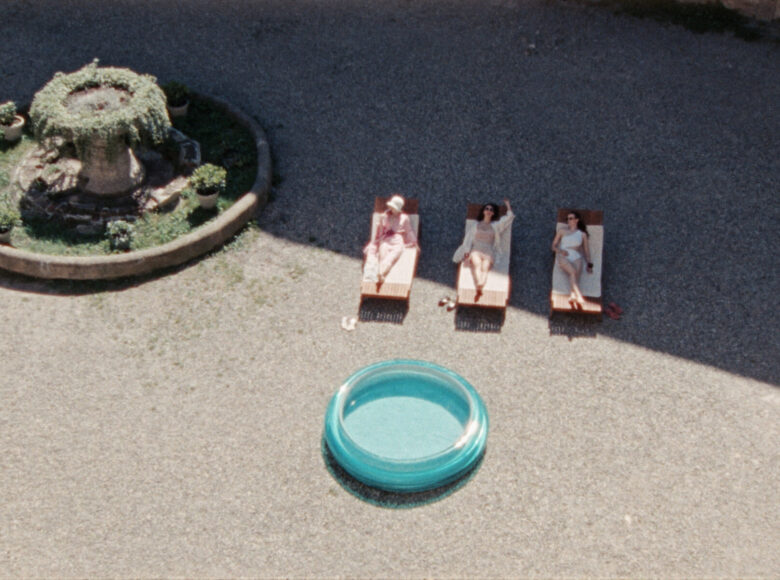 Zdjęcie z lotu ptaka. Przedstawia placyk, na którym na leżakach leżą trzy kobiety w strojach do opalania. Obok kobiet znajduje się klomb z zielonymi roślinami.