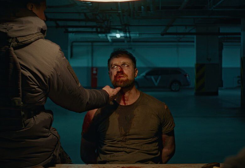 Parking podziemny, półmrok. Na pierwszym planie siedzący mężczyzna z zakrwawioną twarzą. Przed nim, tyłem do nas, stoi mężczyzna w kurtce w kolorze munduru żołnierskiego i przytyka palec dłoni do podbródka siedzącego mężczyzny.