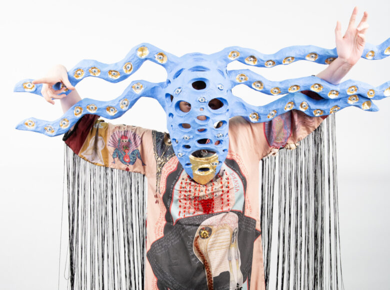 Na białym tle przodem do widza stoi postać w teatralnym stroju: niebieskiej, fantazyjnej masce na twarzy i w kolorowej, szerokiej szacie.