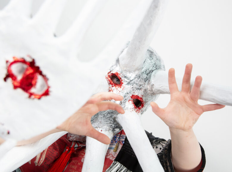 Zbliżenie na kobietę w stroju teatralnym: kolorowej szacie i fantazyjnej masce na twarzy. Kobieta pochyla głowę i unosi dłonie na wysokość swojej twarzy.