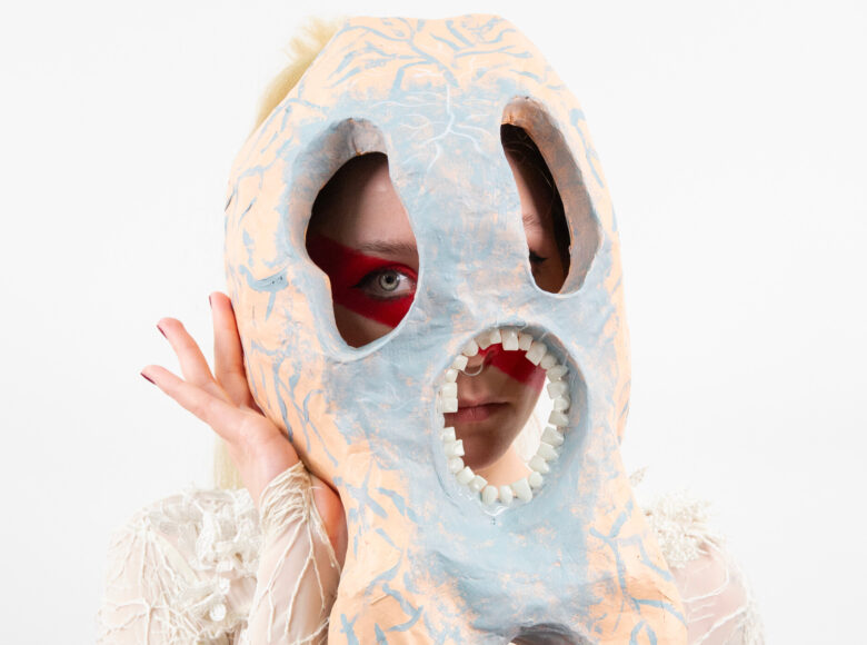 Zbliżenie na osobę w fantazyjnej, teatralnej masce na twarzy. Z tyłu białe tło.