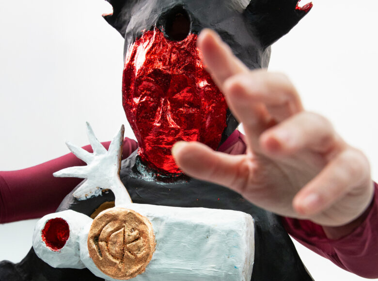 Zbliżenie na osobę w stroju teatralnym: fantazyjnej masce na twarzy oraz bluzce, do której przyczepiona jest figurka, wyglądająca na magiczną lub rytualną. Osoba unosi dłoń do góry. Z tyłu białe tło.