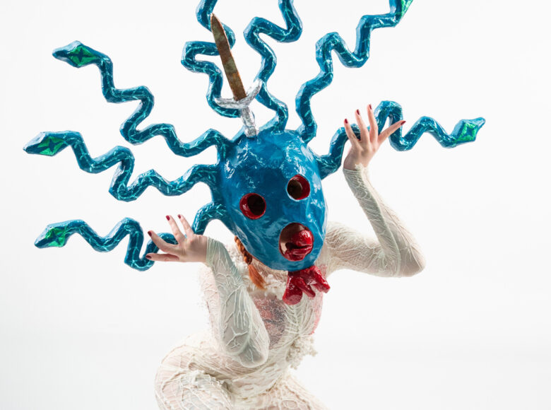 Zbliżenie na kobietę w stroju teatralnym: białym, dekoracyjnym kombinezonie i niebieskiej, fantazyjnej masce na twarzy. Kobieta unosi dłonie na wysokość swojej twarzy. A kobietą białe tło.