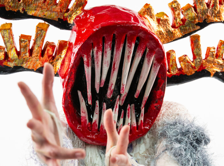 Zbliżenie na osobę w fantazyjnej, teatralnej masce na twarzy. Z tyłu białe tło.