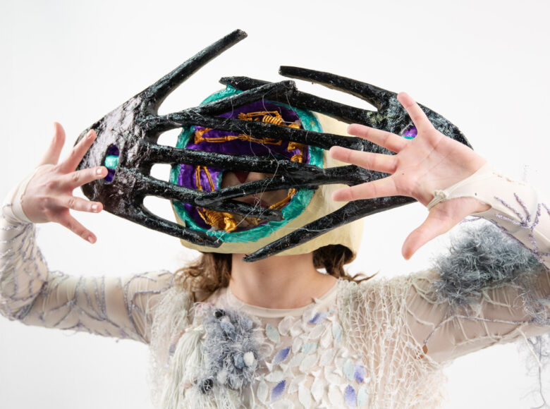 Zbliżenie na kobietę w stroju teatralnym: dekoracyjnej bluzce i fantazyjnej masce na twarzy. Kobieta unosi dłonie na wysokość swojej twarzy.