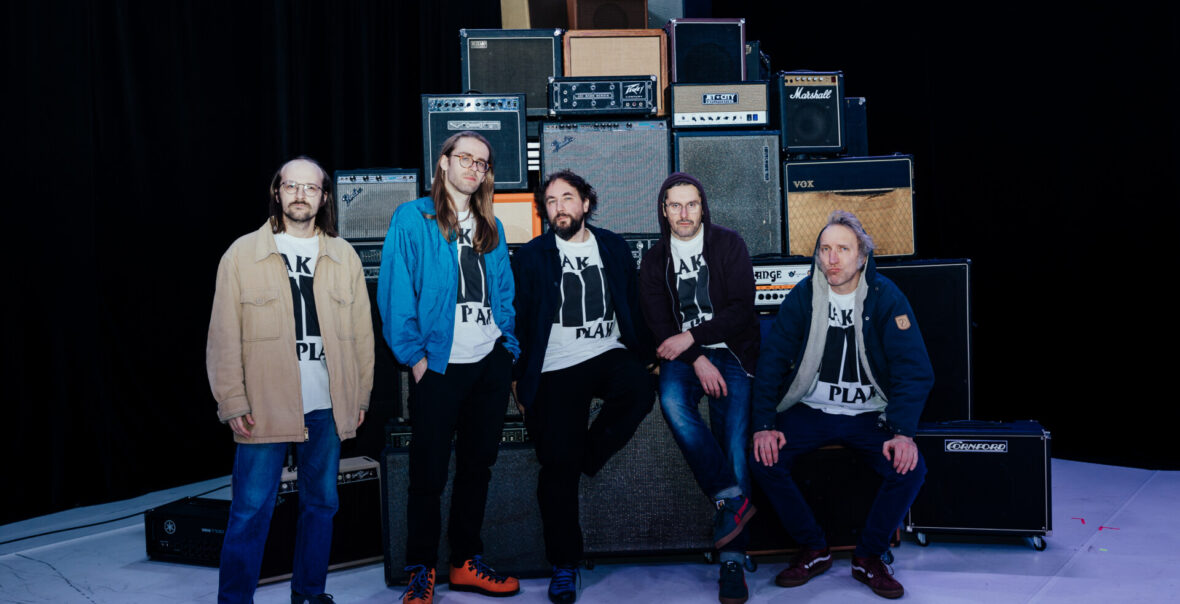 W pomieszczeniu pięciu mężczyzn pozuje na tle głośników, ustawionych w stos. Na ostatnim planie za głośnikami czarne tło.