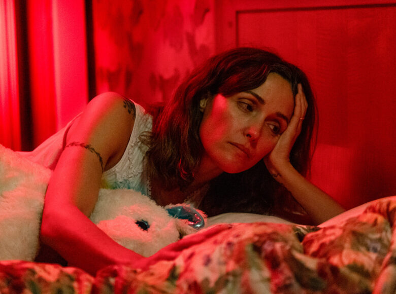 Zbliżenie na kobietę w łóżku, oświetloną czerwonym światłem. Kobieta przybiera pozę półleżącą, obejmując dużą przytulankę i patrząc w bok z zamyślonym wyrazem twarzy.